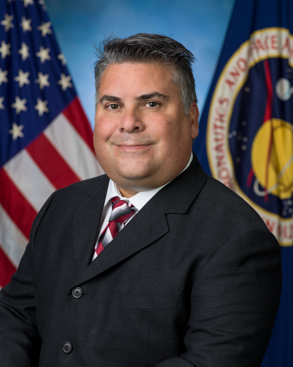 Professional portrait of a Hispanic man wearing a black suit, white shirt, and black and gray striped tie.